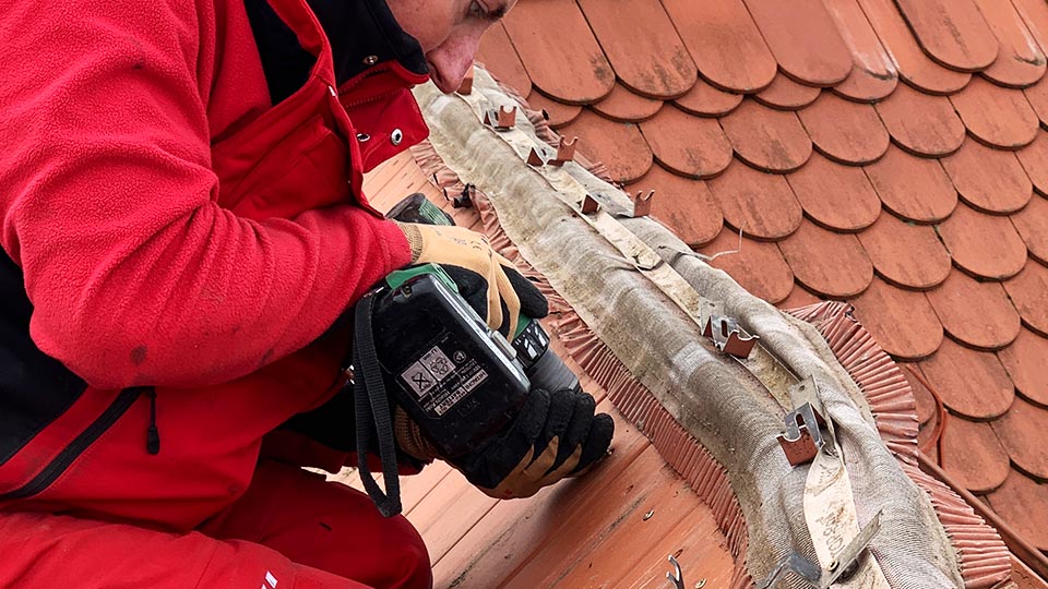 Dachdecker von AMD in roter Jacke setzt mit dem Akkuschrauber Firstklammern an einem Biberschwanzdach Dachdecker von AMD in roter Jacke setzt mit dem Akkuschrauber Firstklammern an einem Biberschwanzdach