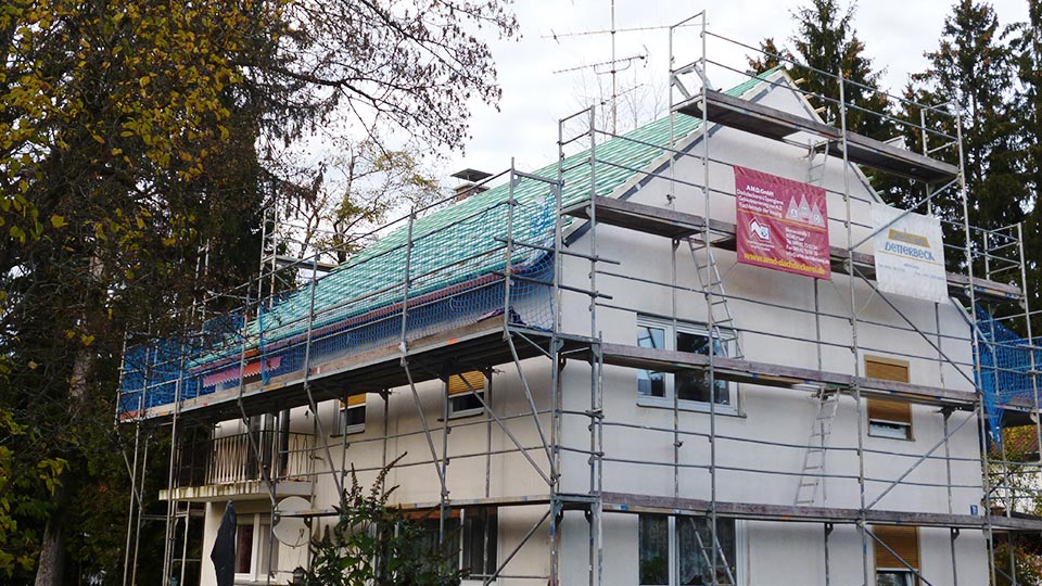 Eingerüstetes Wohnhaus mit Satteldach, auf dem Dach Unterdeckbahn und Lattung, am Gerüst ein Banner von AMD Eingerüstetes Wohnhaus mit Satteldach, auf dem Dach Unterdeckbahn und Lattung, am Gerüst ein Banner von AMD