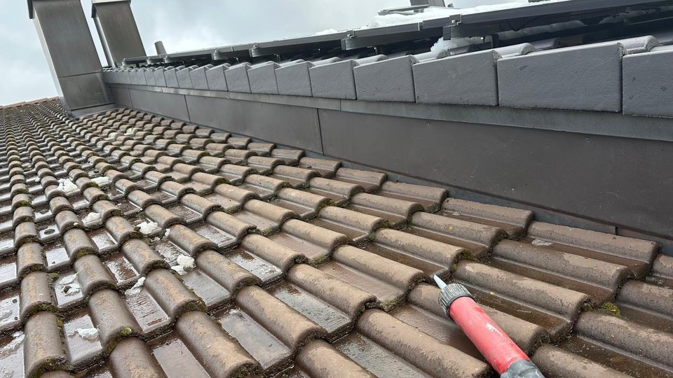 Nasses Ziegeldach mit Ortgangverwahrung aus dunklem Blech, Schnee- und Eisreste auf Ziegeln und Solaranlage, vorne eine Kartuschenpresse Nasses Ziegeldach mit Ortgangverwahrung aus dunklem Blech, Schnee- und Eisreste auf Ziegeln und Solaranlage, vorne eine Kartuschenpresse