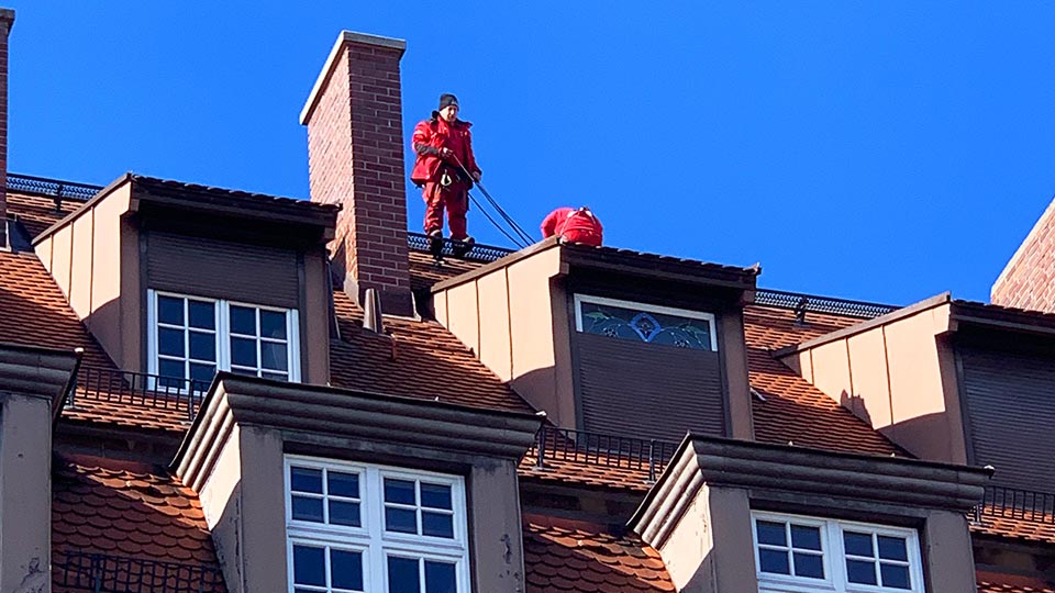Steildach mit Biberschwanzziegeln, Gauben und Ziegelkamin, auf dem First zwei Dachdecker von AMD in roter Arbeitskleidung Steildach mit Biberschwanzziegeln, Gauben und Ziegelkamin, auf dem First zwei Dachdecker von AMD in roter Arbeitskleidung